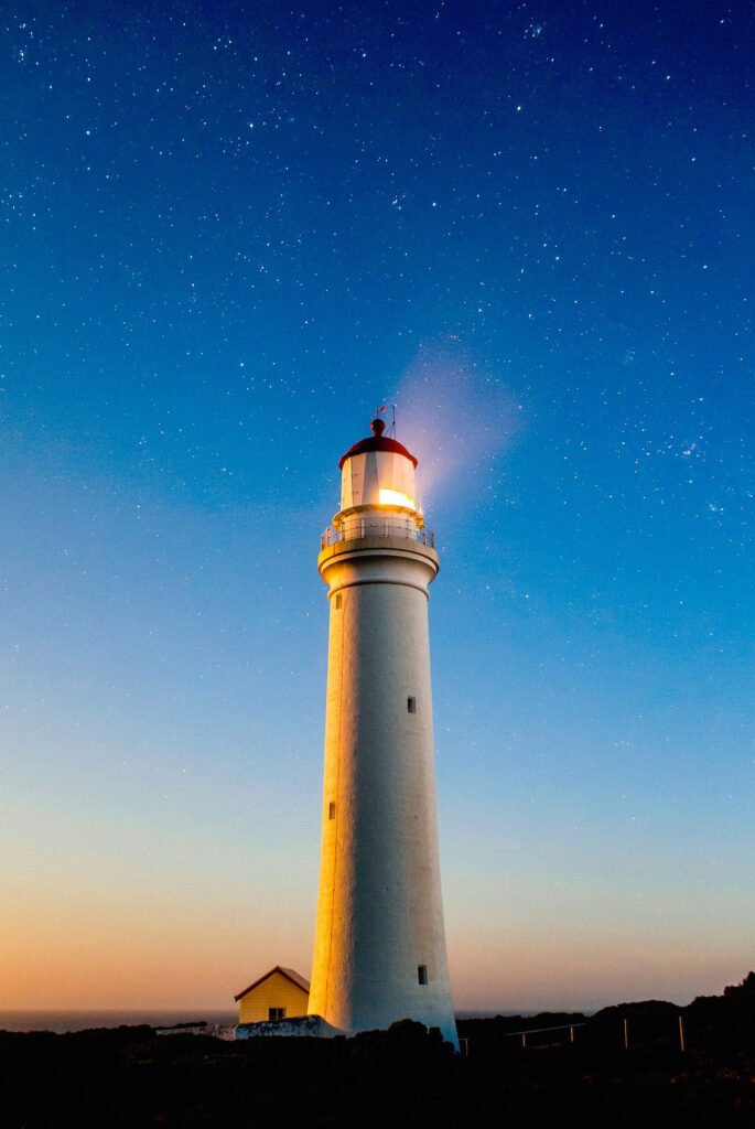 white lighthouse, lit with red roof and small grounds keeper hut on dark land in sky lit by sun set